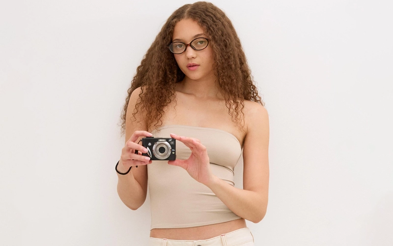 a women having brown curly hair having a camera his hand wearing a skin color Bandeau T-shirts a t-shirt from different types of t-shirts for women