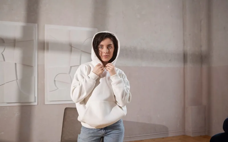 A woman wearing a white hoodie, adjusting the hood while standing indoors in a minimalist setting with light-colored walls