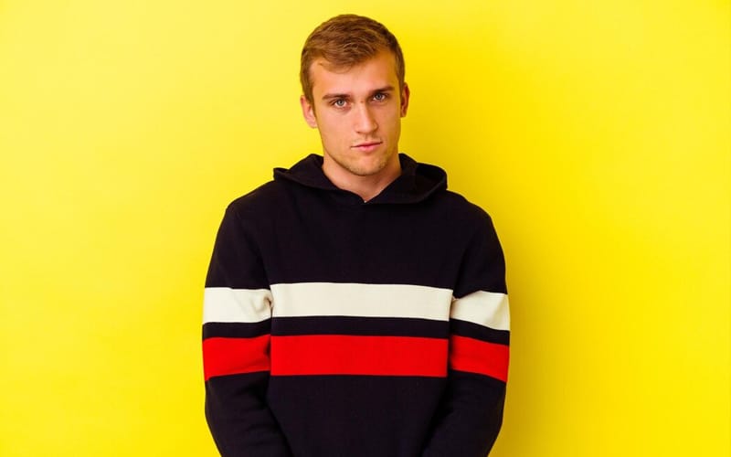 Young man wearing a black hoodie with red, white, and navy blue stripes standing against a yellow background.