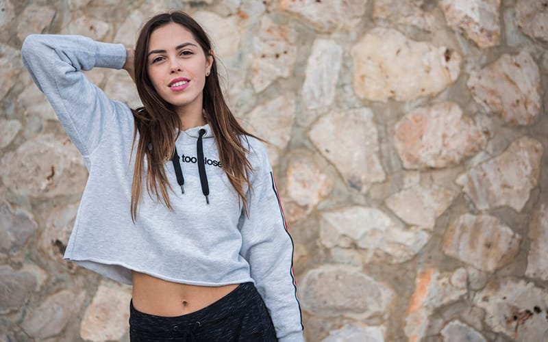 A woman wearing a grey cropped hoodie with ‘you’re too close’ text, standing against a stone wall with one hand behind her head.