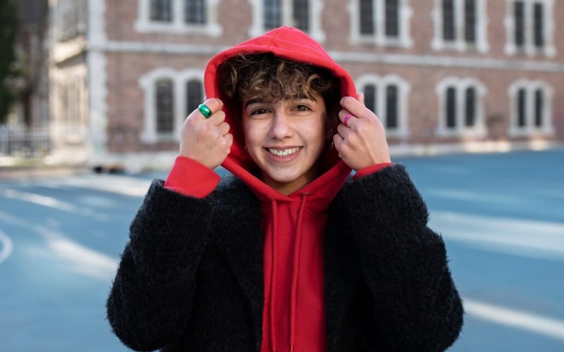 A person smiling and wearing a fleece red hoodie with a black jacket, holding the hoodie’s hood up with both hands, standing outdoors in front of a brick building.