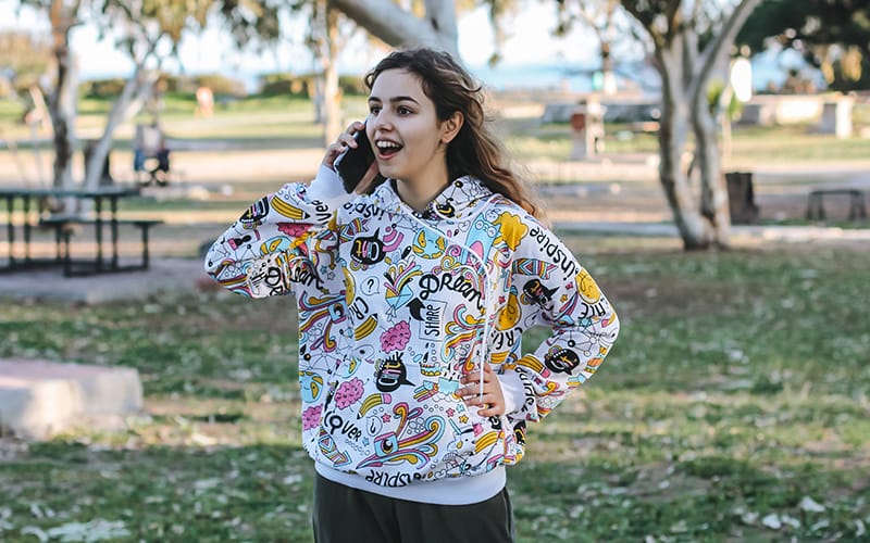 A woman wearing a colorful graphic print hoodie with vibrant patterns, smiling and talking on her phone outdoors in a park.