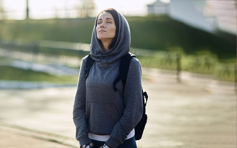 Woman wearing a grey hoodie sweatshirt with a backpack, standing outdoors in natural light.