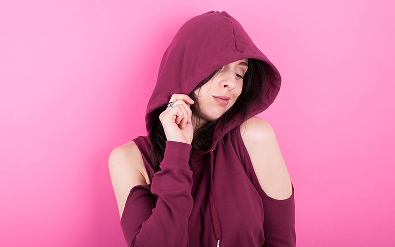 A woman wearing a maroon hoodie with cut-out shoulders, with the hood up, posing against a pink background.