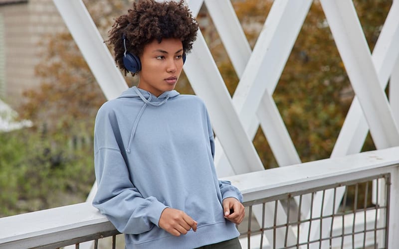 A young woman wearing a light blue hoodie with a hood, standing outdoors with headphones on, resting her arm on a metal railing.
