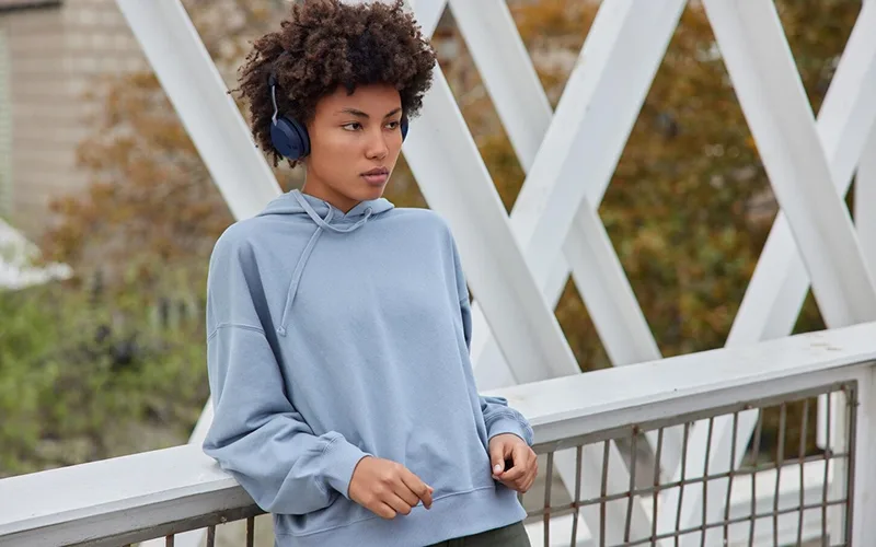 A young woman wearing a light blue hoodie with a hood, standing outdoors with headphones on, resting her arm on a metal railing.