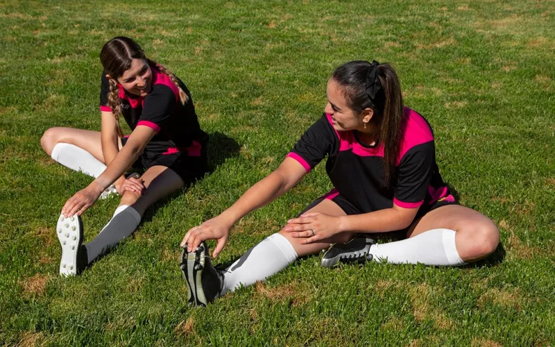 Two women stretching before a soccer game, wearing black and pink jerseys with soccer socks.