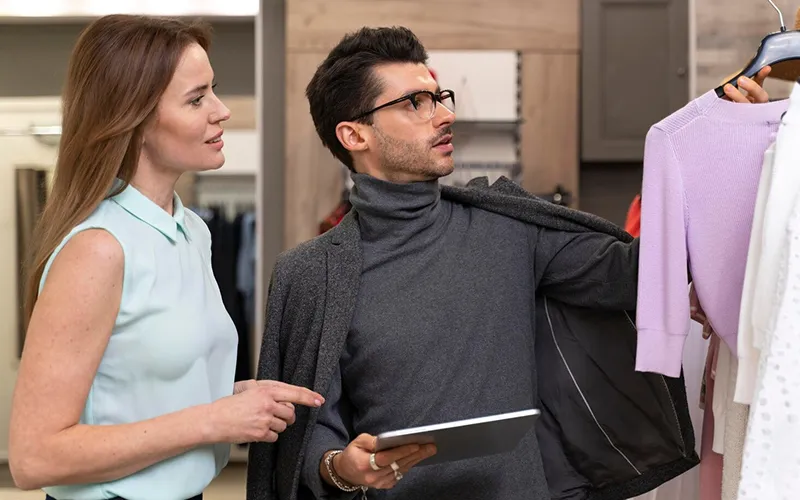 A woman and man discussing clothing at a fashion store, with the man holding a tablet and pointing to a shirt on a rack.