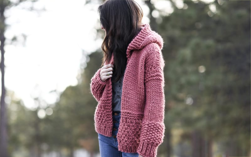 A girl wearing a cozy knitted pink hoodie, standing outdoors with a blurred natural background.