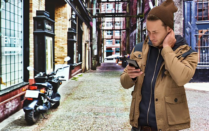 A man wearing a beige motorcycle hoodie, standing outdoors in a city alley, looking at his phone with a relaxed posture, a motorcycle in the background.
