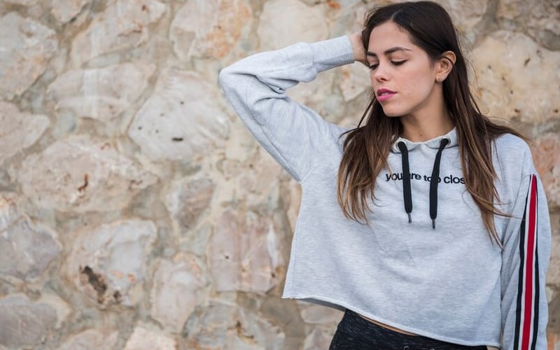 A girl wearing a grey off-shoulder hoodie with a ‘you’re too close’ text design, standing against a stone wall, with a relaxed pose
