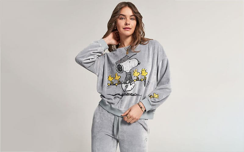 Woman wearing a grey Snoopy sweatshirt with a cartoon design featuring Snoopy and Woodstock, standing and smiling.