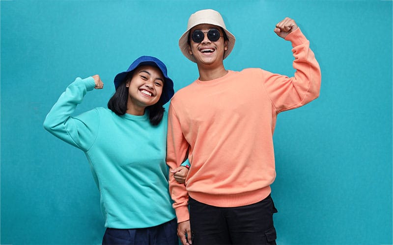 Smiling man and woman wearing colorful unisex sweatshirts, posing with raised fists in front of a teal background.