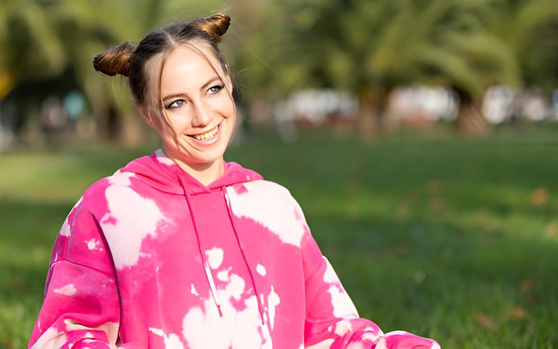 A woman wearing a bright pink tie-dye hoodie with a smiling expression, sitting outdoors with her hair styled in two buns