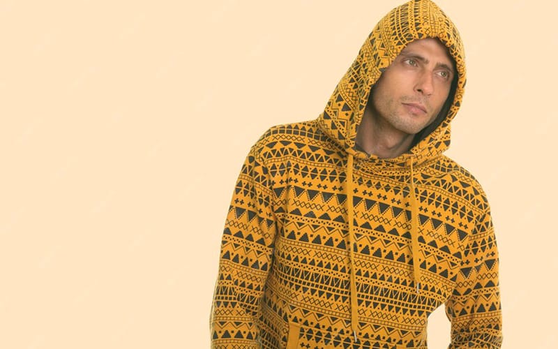 A man wearing a yellow waffle knit hoodie with a tribal geometric pattern, standing with the hood up against a soft beige background.