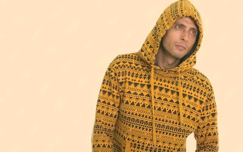 A man wearing a yellow waffle knit hoodie with a tribal geometric pattern, standing with the hood up against a soft beige background.