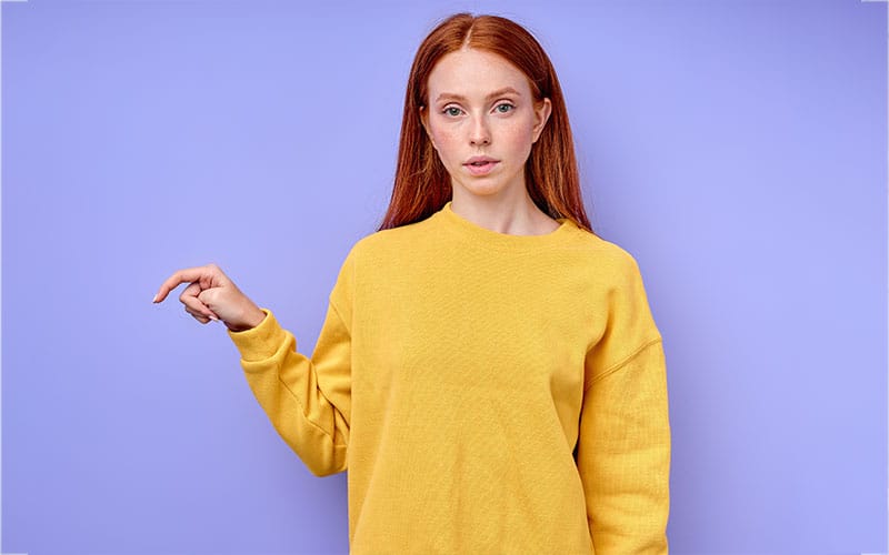 Woman wearing a yellow sweatshirt, pointing at something, with a purple background.