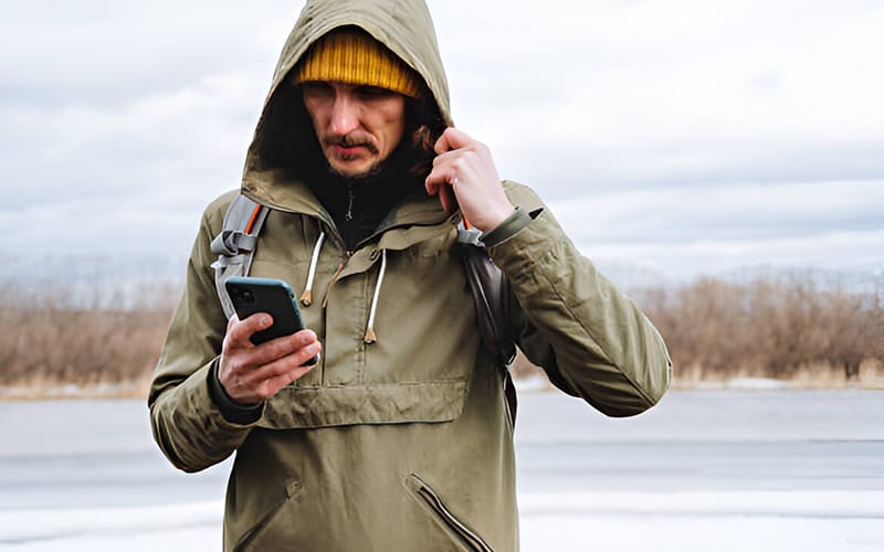 A man wearing a green windbreaker hoodie and a yellow beanie, standing outdoors in a wintery setting, looking at his phone with a backpack on