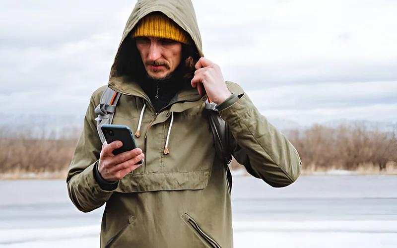A man wearing a green windbreaker hoodie and a yellow beanie, standing outdoors in a wintery setting, looking at his phone with a backpack on