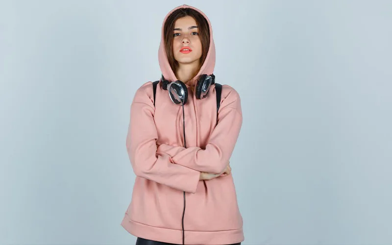 Young woman wearing a pink zipper hoodie with headphones around her neck, standing against a light blue background.