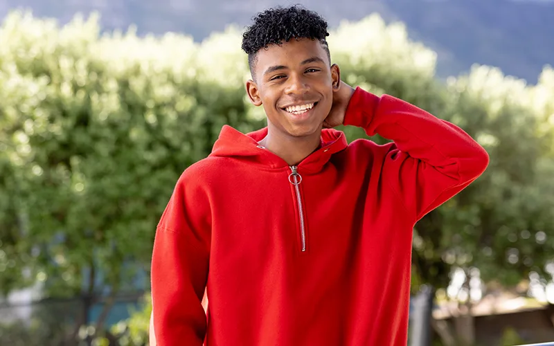 A smiling young man wearing a red half-zip hoodie, standing outdoors with greenery in the background.