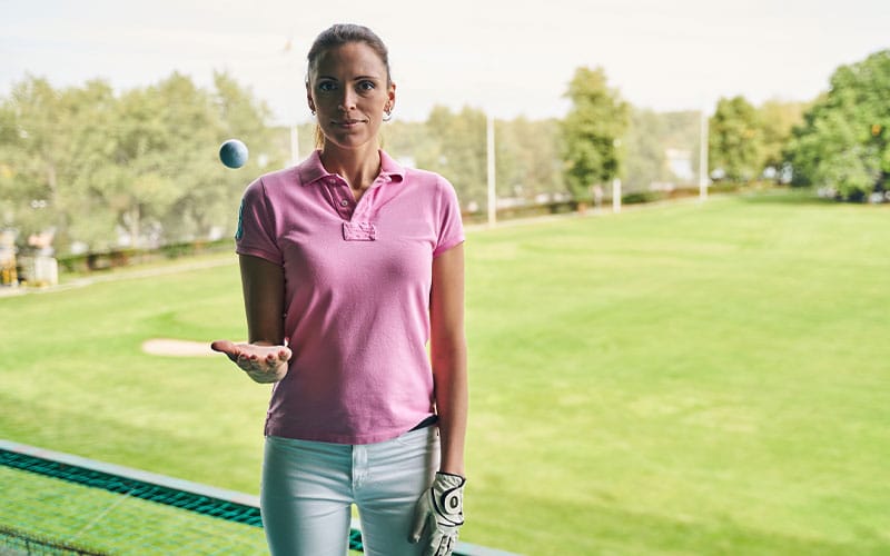 Woman wearing a pink polo shirt and golf gloves, holding a golf ball in her hand, standing on a golf course