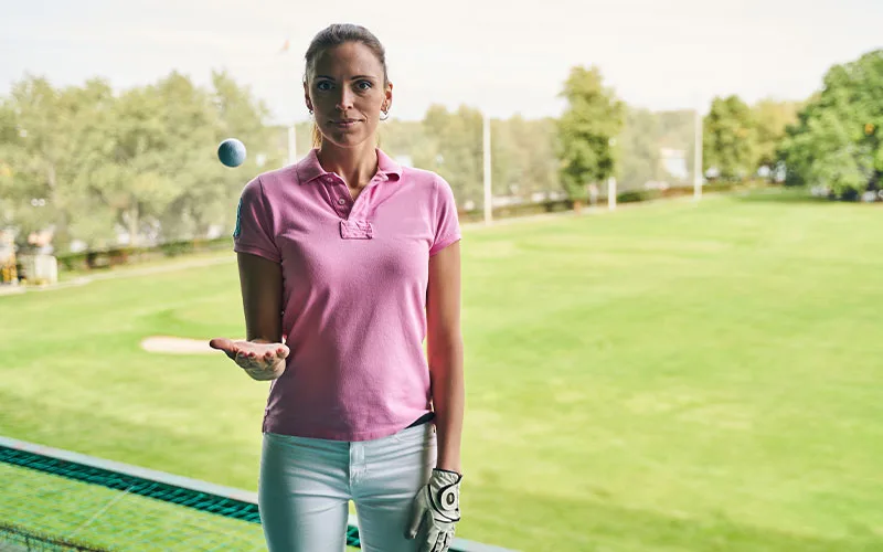 Woman wearing a pink polo shirt and golf gloves, holding a golf ball in her hand, standing on a golf course