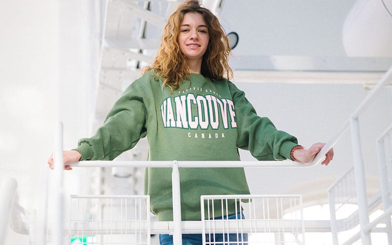Woman wearing a vintage green sweatshirt with 'Vancouver Canada' printed on it, standing on a balcony in a modern, bright setting