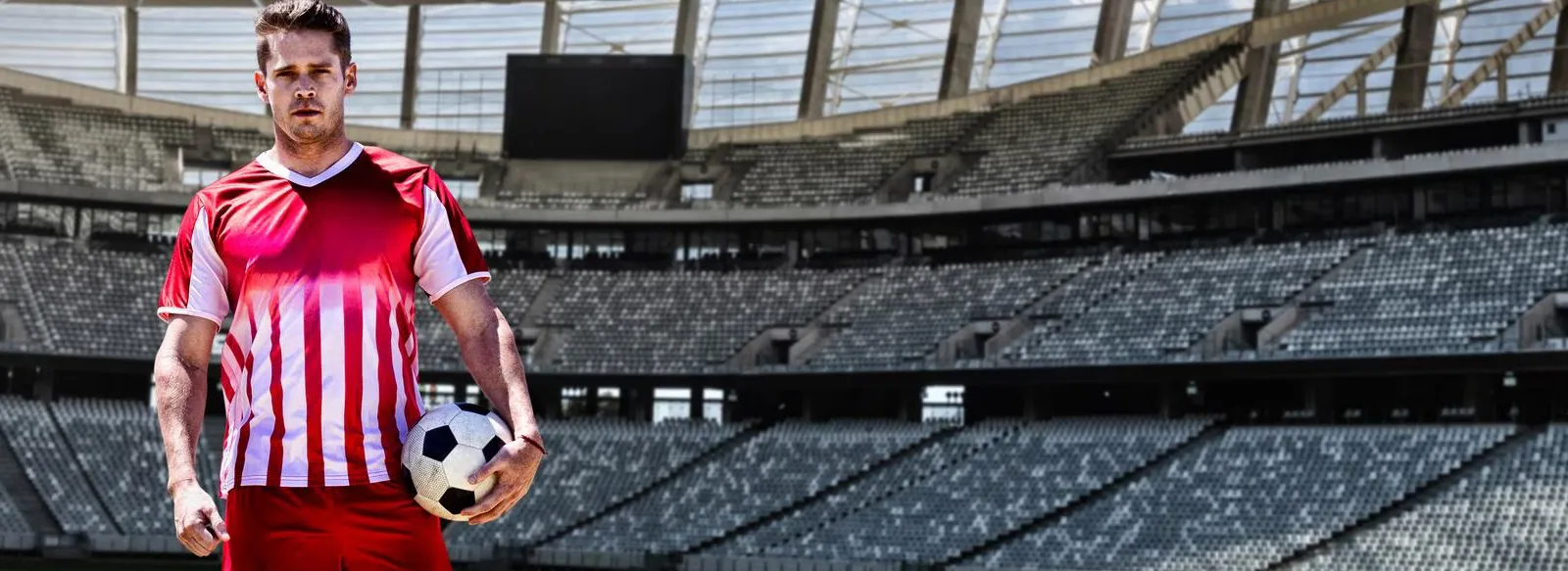 A man standing in a large soccer stadium, holding a soccer ball while wearing a red and white striped jersey