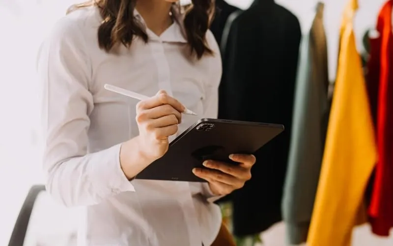 A person in a white shirt using a digital stylus to work on a tablet in a clothing studio, with colorful garments hanging in the background.