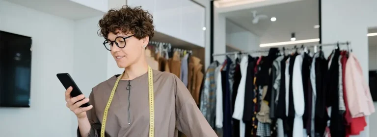 A fashion designer holding a phone while wearing a measuring tape in a well-lit clothing shop, surrounded by racks of clothes