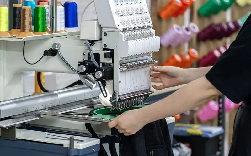  A close-up of an embroidery machine in use, with a person carefully adjusting fabric for custom embroidery. Colorful spools of thread are visible in the background, showcasing the precision and craft involved in the embroidery process.