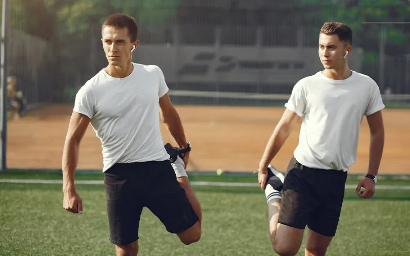 Two young men stretching before their soccer game on a field, both wearing white t-shirts and black shorts.